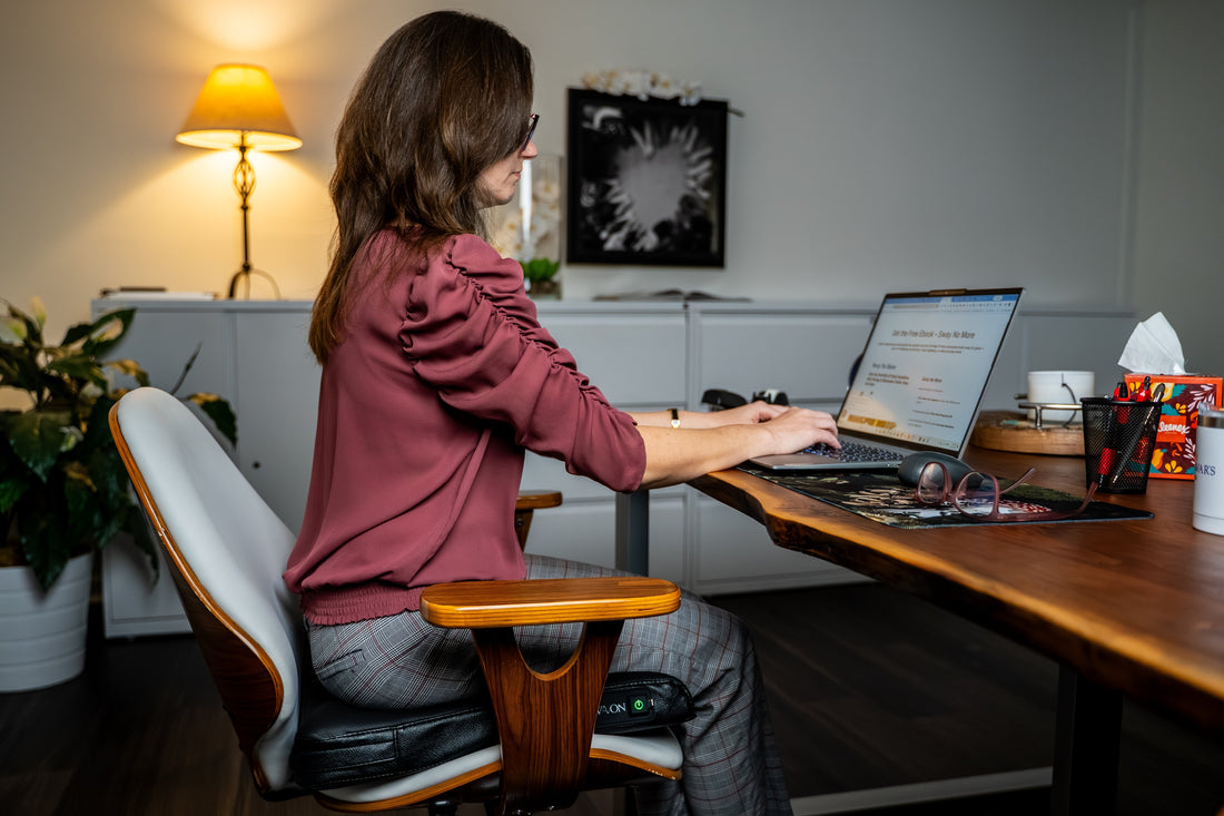 A woman working on a laptop while sitting on a black Wavon ergonomic cushion for passive wellness at a wooden desk.